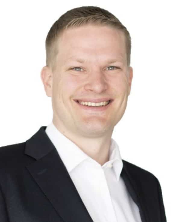 Ein lächelnder Mann in dunkler Anzugsjacke und weißem Hemd, mit kurzen hellen Haaren, posiert vor einem schlichten weißen Hintergrund. Sparkpoint Executive Coaching.